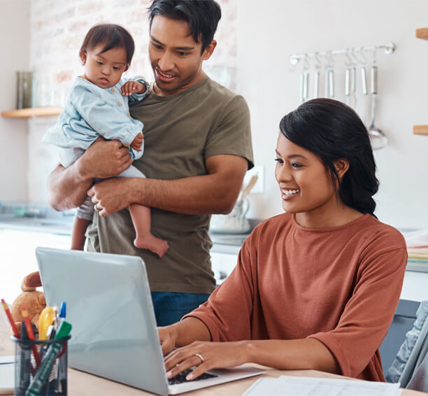 Father holds baby as mother uses laptop for financial planning