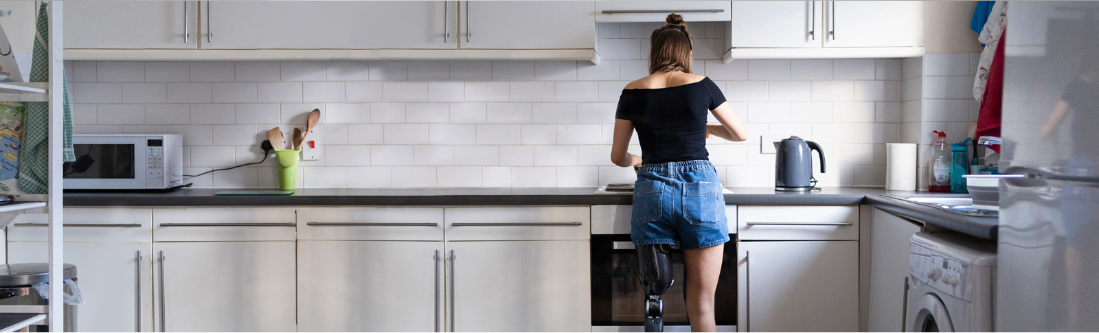 Woman standing at her kitchen stove, her back is to the camera. She has a full prosthetic left leg. She is wearing a black short-sleeved shirt and jean shorts. The tidy kitchen has black counters, white cabinets and white wall tiles.