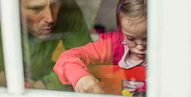 Young girl with Down Syndrome wearing glasses and a pink hooded sweatshirt playing with her father.